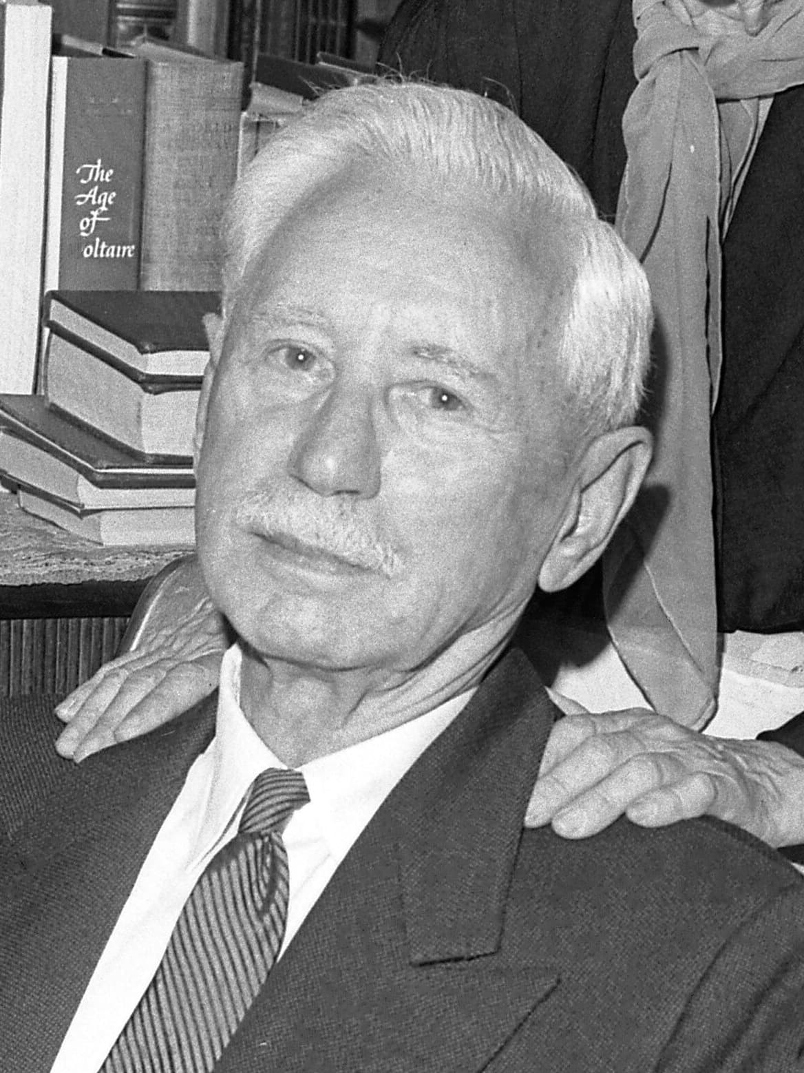 Black-and-white photograph of Will Durant, white-haired with a moustache, in a suit and tie, seated before a shelf of books.